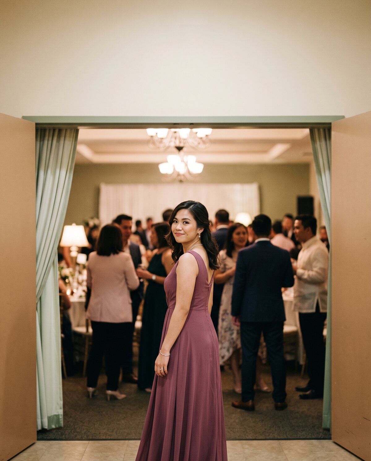 A woman in a berry-colored gown pausing at the entrance of an event hall, glancing back with a smile as guests mingle behind her.